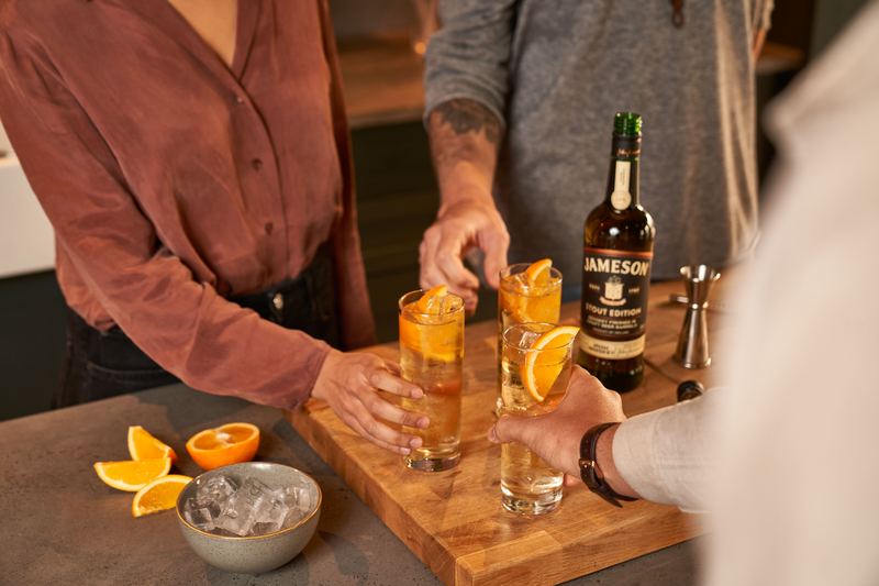 Friends toasting with whiskey cocktails garnished with orange slices, featuring Jameson Stout Edition on a wooden cutting board with fresh orange wedges and ice, creating a warm and inviting atmosphere.