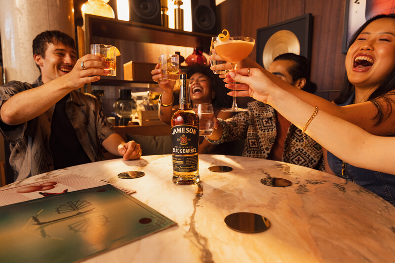 A group of friends joyfully toasting with whiskey cocktails around a stylish marble table, featuring a bottle of Jameson Black Barrel, creating a warm and celebratory atmosphere in a chic lounge setting.