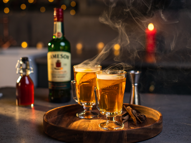 Tow steaming hot whiskey cocktails served in elegant glass mugs on a wooden tray, accompanied by cinnamon sticks and a bottle of Jameson Irish Whiskey, set in a cozy, candlelit ambiance.