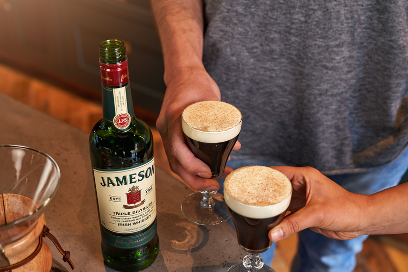 Two Irish Coffee cocktails topped with frothy cream and a dusting of spice, served alongside a bottle of Jameson Irish Whiskey on a cozy kitchen counter, creating a warm and inviting atmosphere.