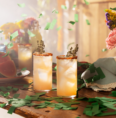 Two refreshing whiskey cocktails served in tall glasses with ice, a spiced rim, and a sprig garnish, set on a festive wooden table with green confetti, fresh flowers, and warm sunlight.