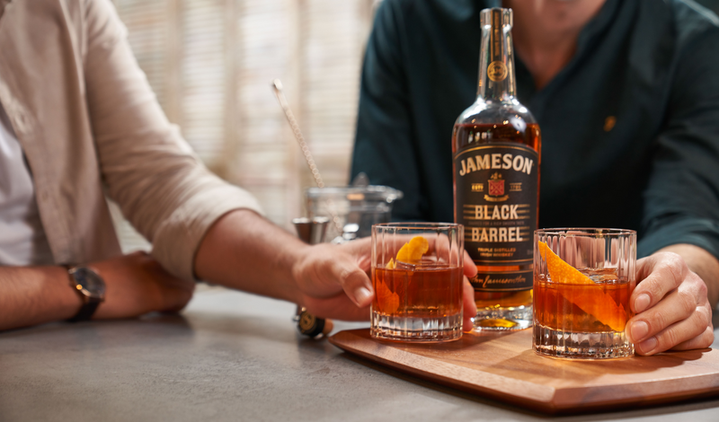 Two friends enjoying whiskey old fashioned cocktails garnished with orange peels, featuring a bottle of Jameson Black Barrel on a wooden tray, creating a refined and inviting atmosphere.