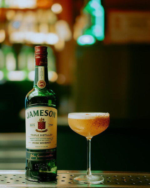 A bottle of Jameson Irish Whiskey beside a coupe glass with a golden cocktail, frothy top, and spiced rim, set against a warmly lit bar background.