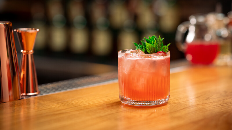 A vibrant pink cocktail served over ice in a textured glass, garnished with fresh mint and a strawberry, set on a polished wooden bar with copper bar tools and a blurred background.